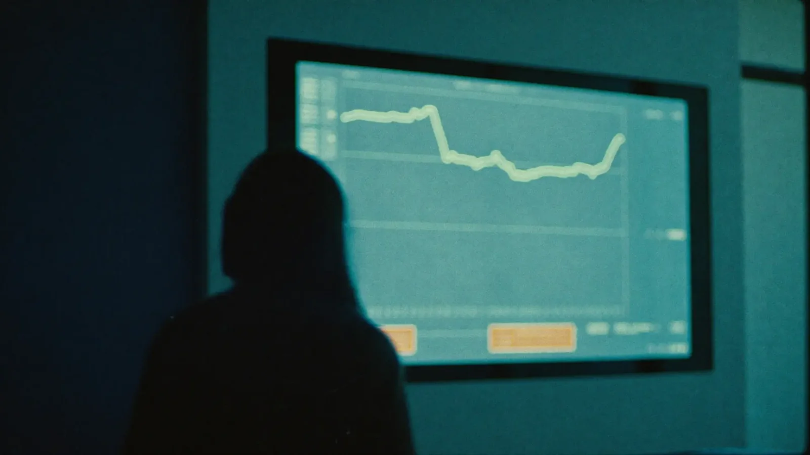 Silhouette of a person in a dim room looking at a glowing monitor showing a data line graph